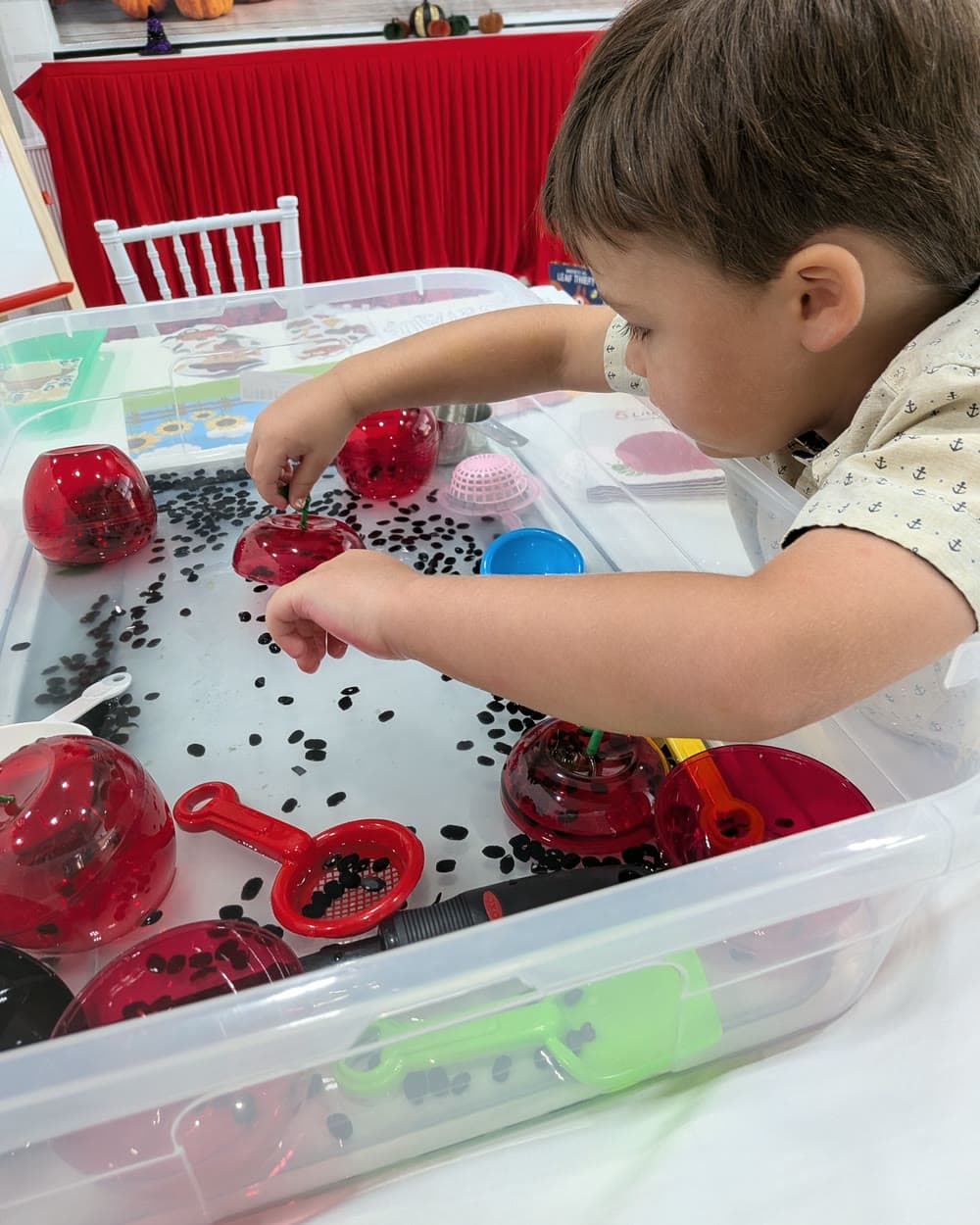 A boy reaches into a water bin of red toy apples, black beans and scoops