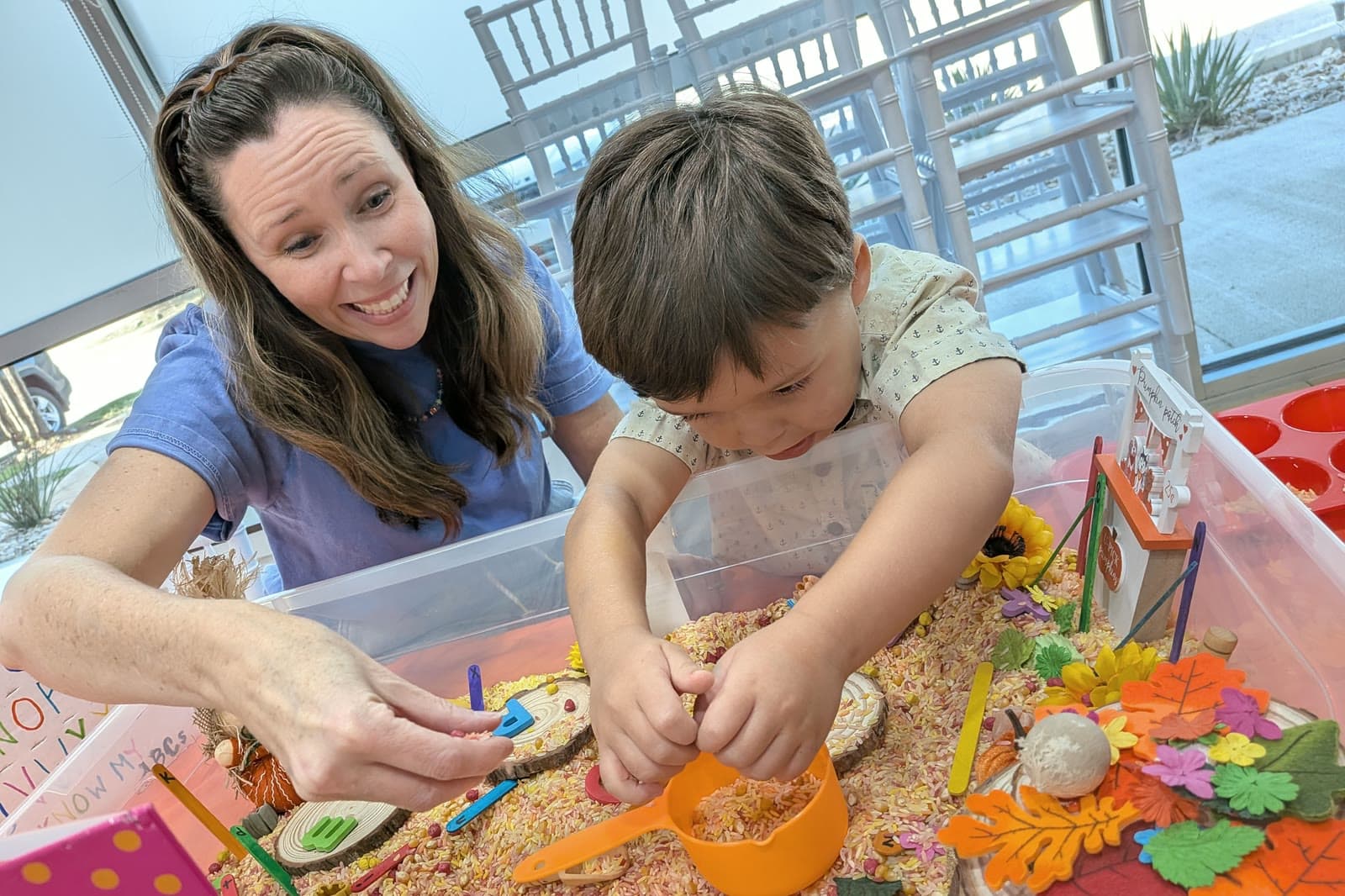 Ms. Nora laughs as a boy pours colored rice into an orange cup in a fall sensory bin