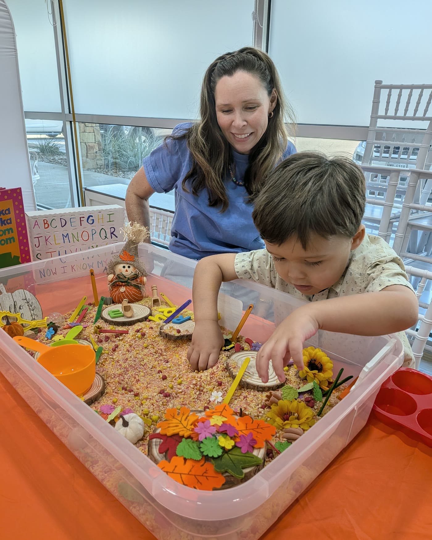 Ms. Nora smiles beside a boy who digs both hands into a fall sensory bin of colored rice, leaves and sunflowers