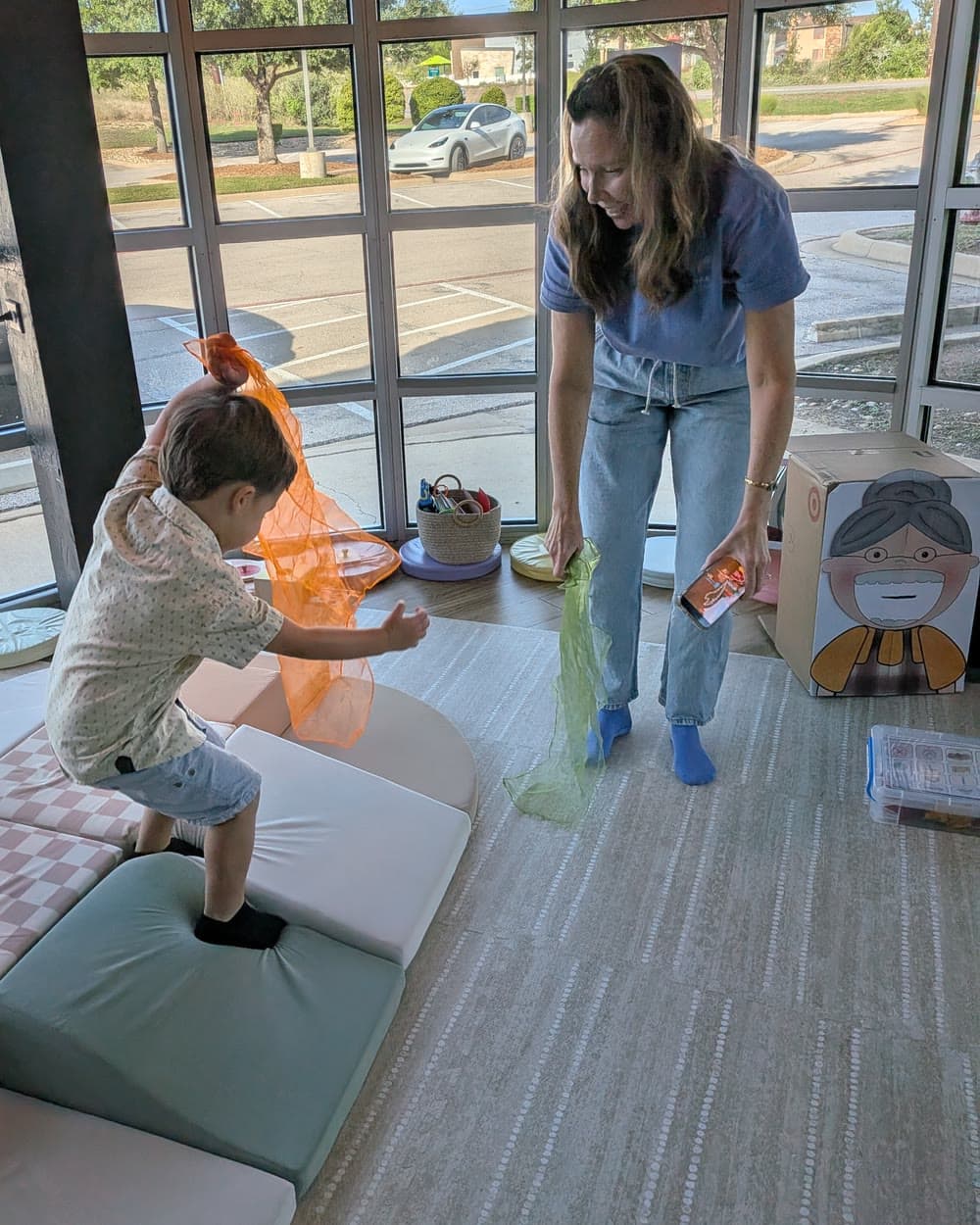 A boy on soft play blocks waves an orange scarf while Ms. Nora dances with a green one