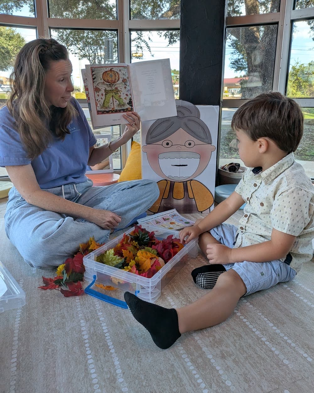 Ms. Nora holds a picture book open for a boy, with a bin of fall leaves between them