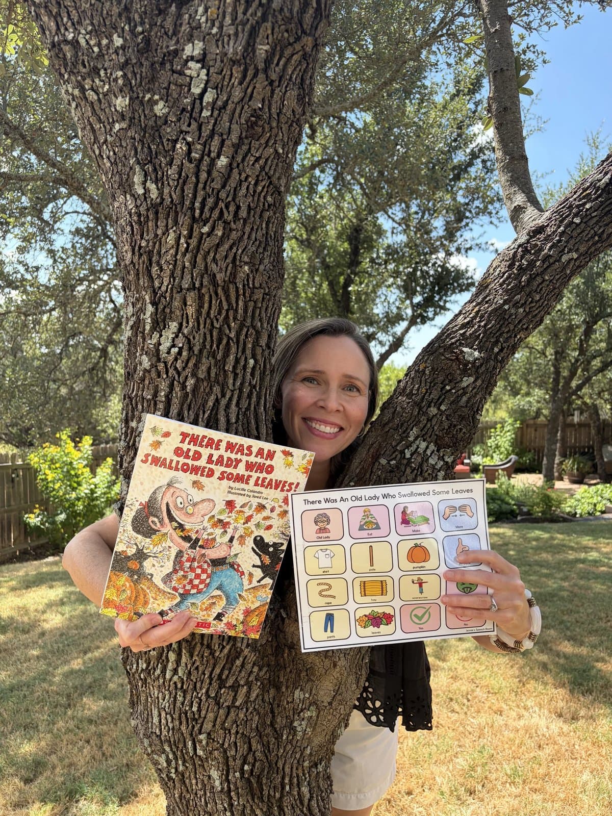 Ms. Nora smiles under a tree, holding a picture book and a picture board for the story