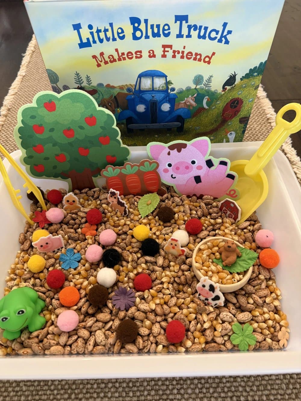 A farm sensory bin of dried corn and beans with pompoms, a pig, a cow, and a frog, set in front of Little Blue Truck Makes a Friend