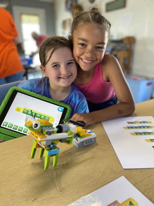 Two girls grin beside the brick robot they built and the tablet that runs it