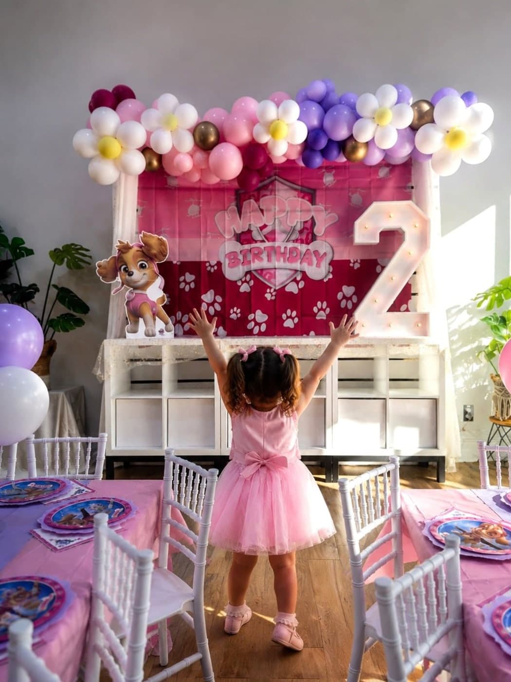 A little girl in a pink tutu with her arms raised in front of her birthday backdrop and balloon garland
