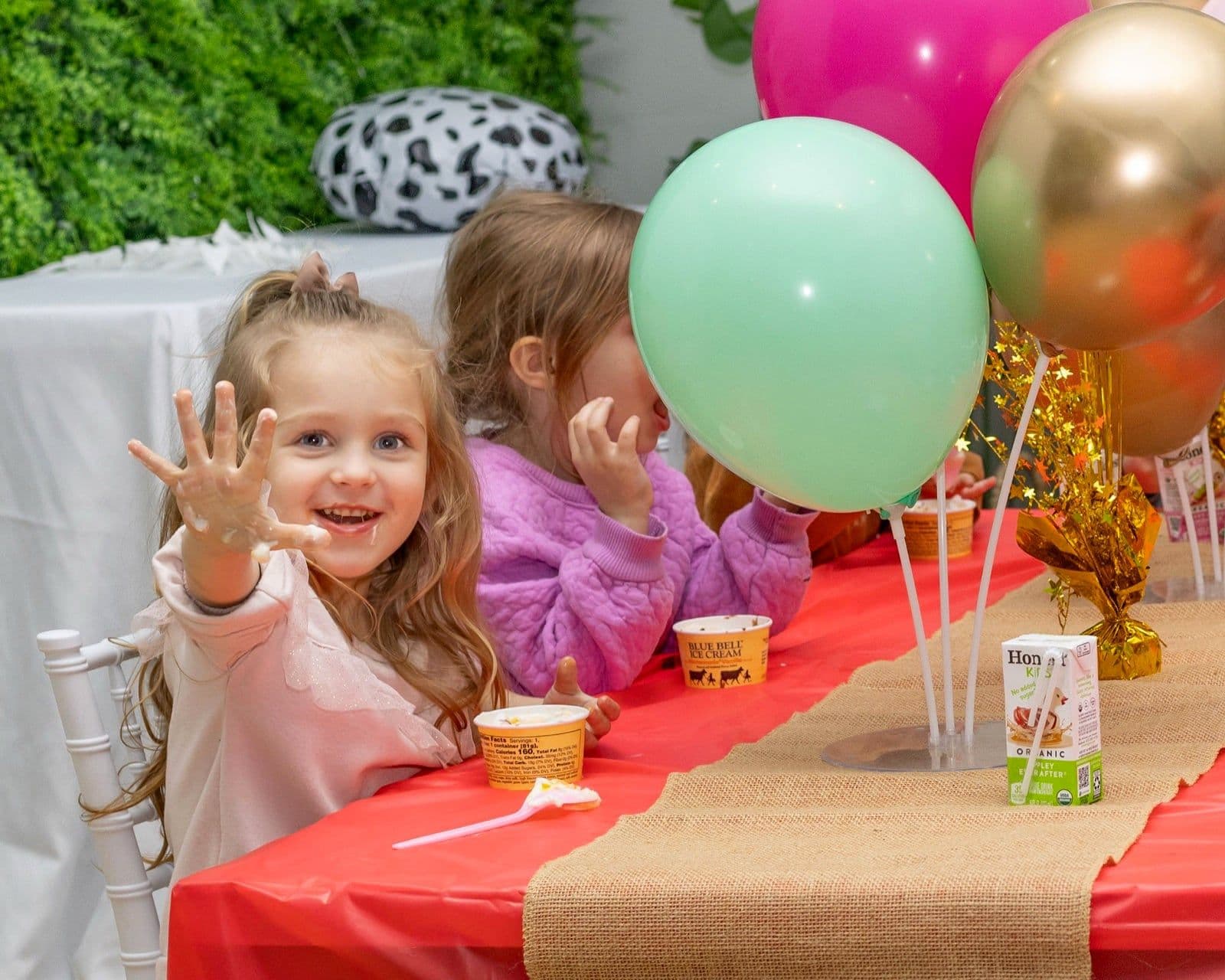 Two children eating ice cream at a decorated party table under balloons