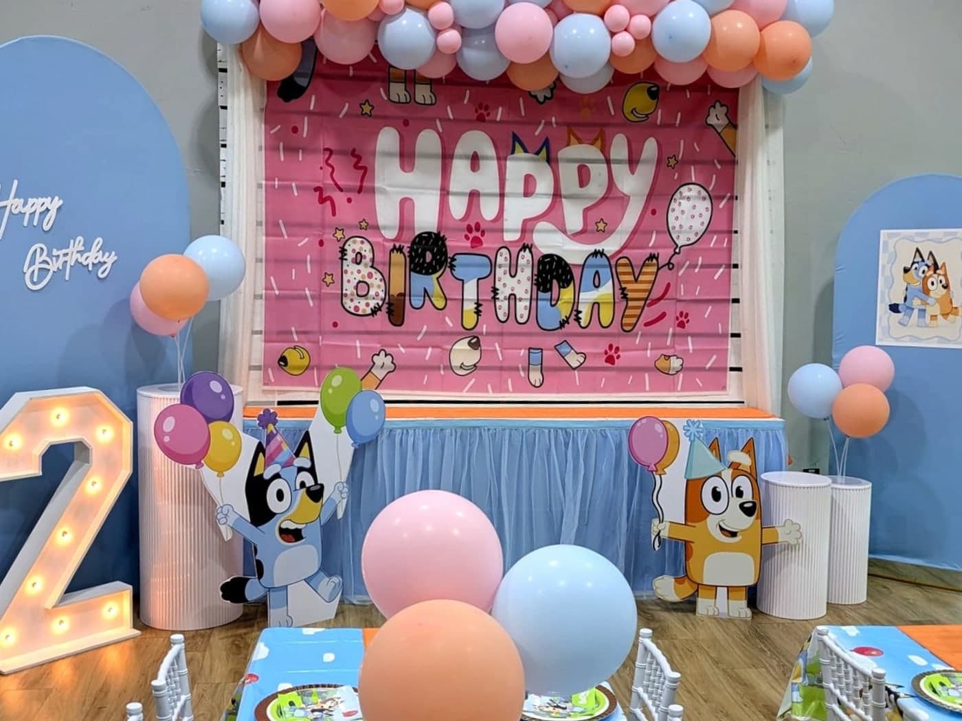 Blue and peach setup with a pastel balloon garland, a pink birthday backdrop, cartoon dog cutouts and a lit marquee number two