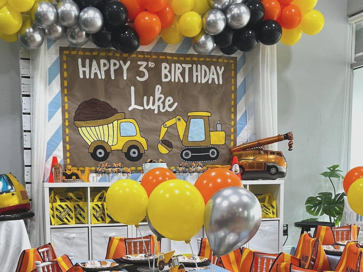 Construction setup with a hand painted digger backdrop, a yellow and black balloon garland and hi-vis vests on the chairs