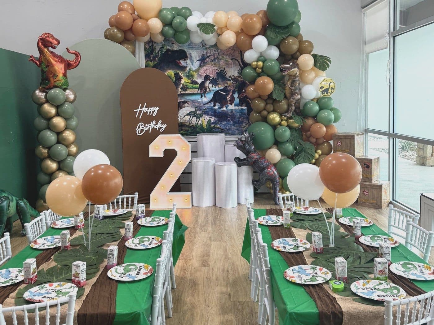 Dinosaur setup with a sage and gold balloon arch, a prehistoric backdrop and a lit marquee number