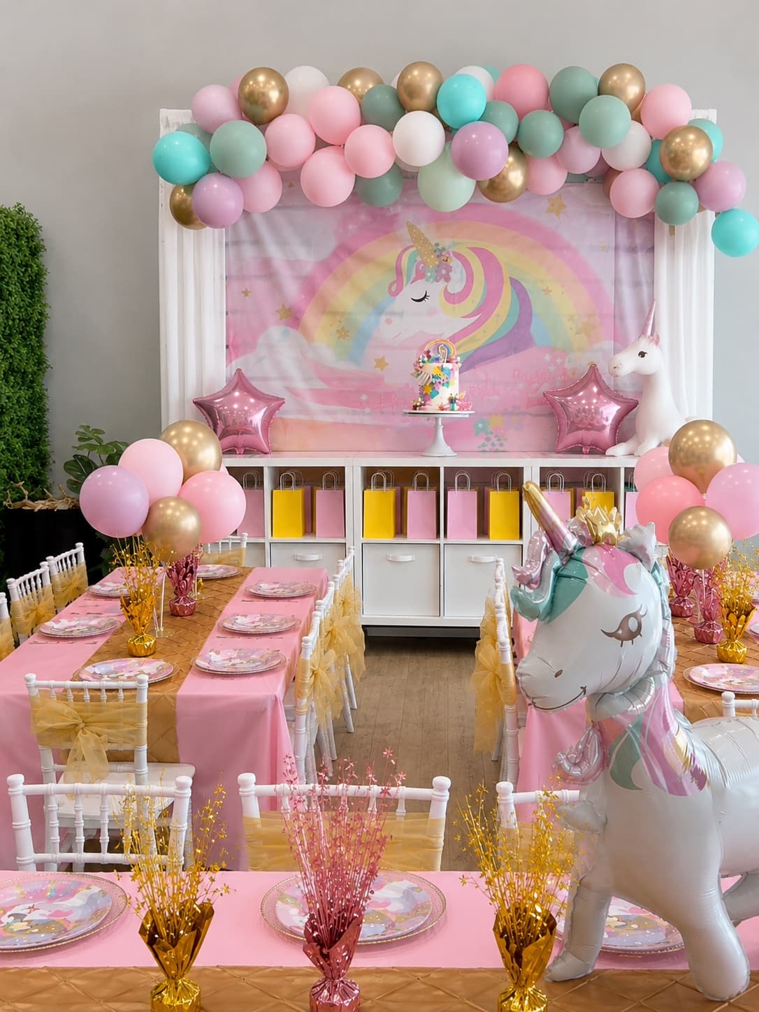 Pastel unicorn birthday party setup with balloon garland, cake, and favor bags