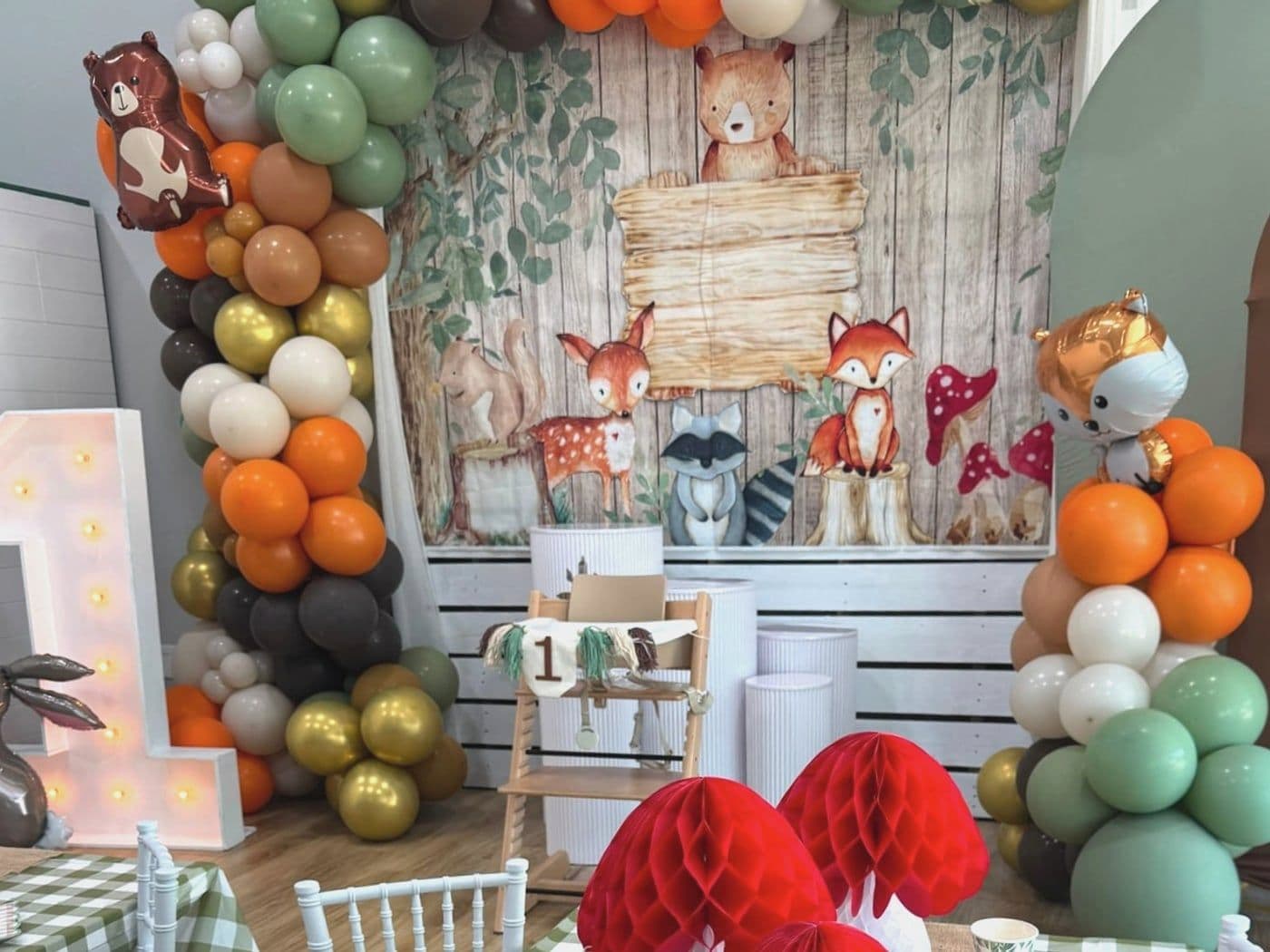 Woodland setup with a forest animal backdrop, a sage and rust balloon arch, paper mushrooms and gingham tables
