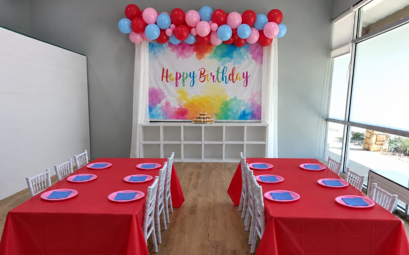 Two long tables set with red tablecloths, pink plates and blue napkins, facing a rainbow Happy Birthday backdrop under a balloon garland